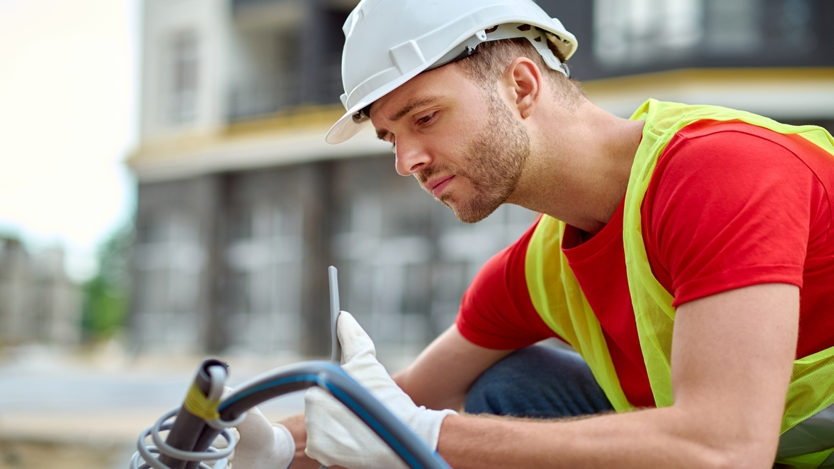 Adjustment. Young adult man in protective helmet gloves and bright vest crouching looking at tool sideways to camera at construction site